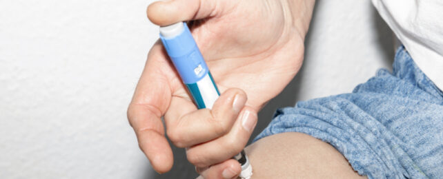 Close-up of person's hand holding insulin injection pen on upper thigh.