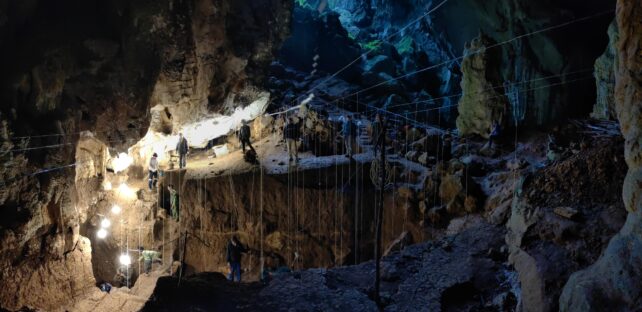 Researchers standing on edge of excavated pit, with lamps lighting the dark cave and tape measures strung across the site.