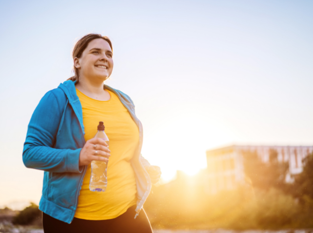 A smiling woman with brown hair in a ponytail is wearing a yellow top with a blue jacket is holding a water bottle while running outdoors at sunrise. 