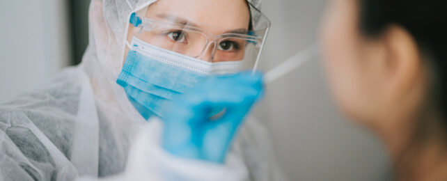 Healthcare worker wearing a blue surgical mask and white disposable gown taking nasal swab from a patient.