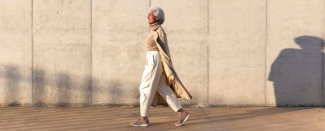 Grey-haired woman wearing white pants, beige trenchcoat and sneakers, walking alone in front of cement wall.
