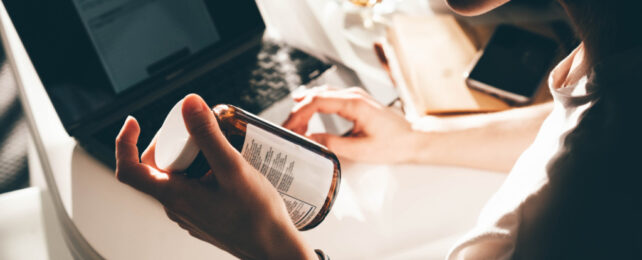 Close-up of person holding medicine bottle in left hand, right hand on keyboard of open laptop.