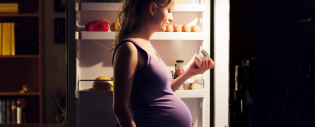 Pregnant woman wearing purple top holding yoghurt tub in front of open fridge, at night.