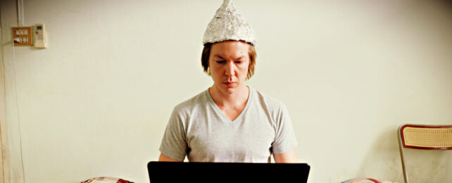 Man wearing tin foil hat looks at laptop
