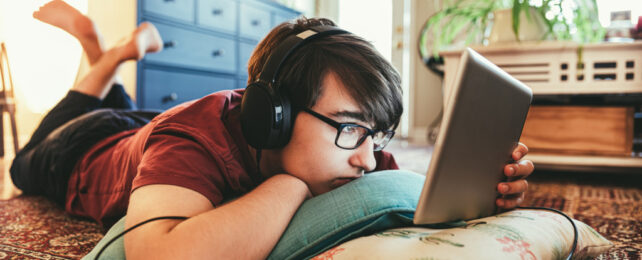 Teenage boy with glasses reading from tablet, lying on floor, wearing headphones.