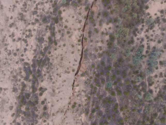 Aerial view of long crack in bare ground running through sparse scrubland.