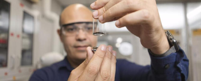 Bald man wearing safety glasses in lab hold glass vial containing black material.
