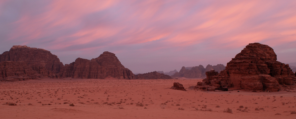 Red tinged desert landscape with rocky outcrops and sunrise stained clouds above