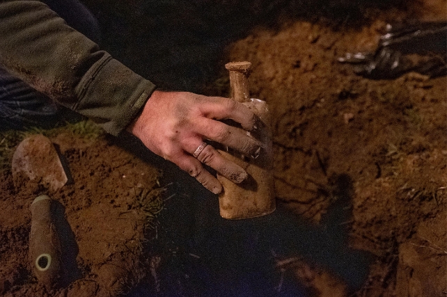 An outstretched arm holding a dirty bottle, with a spade and dirt in background