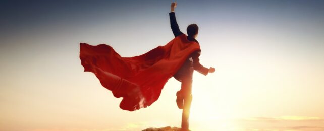 A person standing on a mountain top with fist in the air wearing a red cape