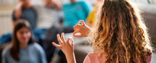 Girl with curly blonde hair standing in front of group sitting on couch, playing charades.