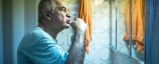 Grey-haired man brushing teeth in front of bathroom mirror, in low lighting and with orange towel hanging on wall.