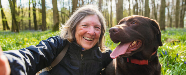 Grey-haired woman takes selfie with brown dog in forest.