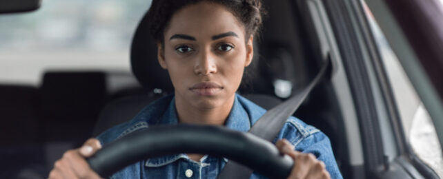 Young black woman wearing blue denim shirt holding steering wheel, with blank facial expression.