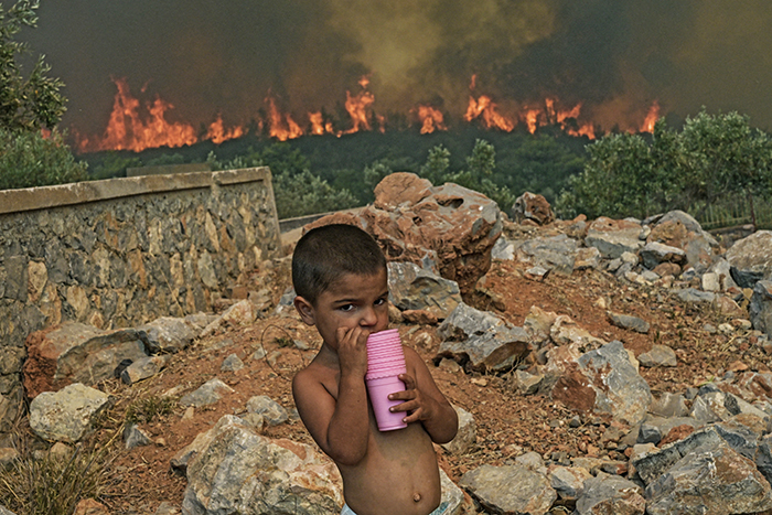 Child taking a drink in his hard as fires blaze in the background