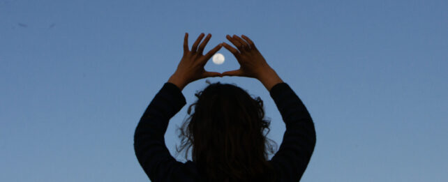 the full moon in a clear crepuscular light being appreciated and framed between index fingers and thumbs by a teenager in silhouette
