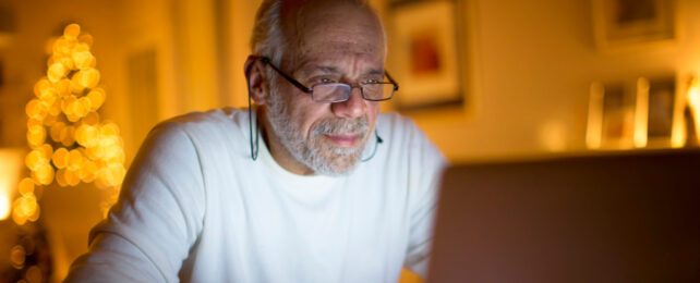 Grey haired man wearing reading glasses working on laptop in low, yellow light.