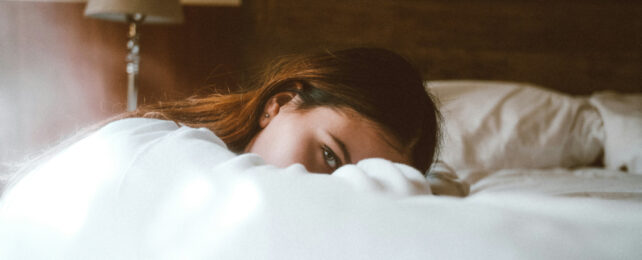 Brown haired woman with forehead resting crossed arms lying on bed, one eye looking directly at camera.