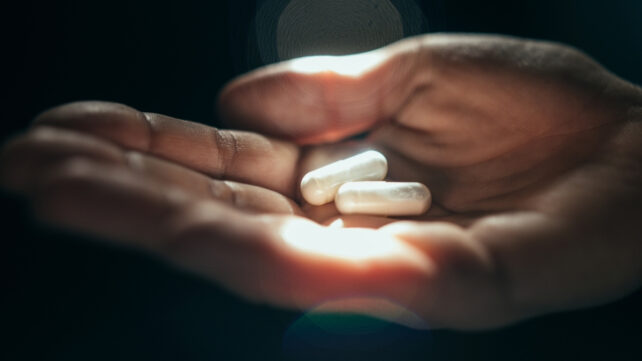 Close up of a hand holding 2 white capsule pills in sunlight, black background