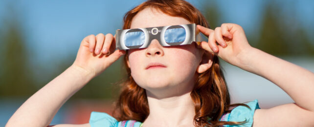 Red haired girl wearing solar eclipse glasses on a blue-sky day.