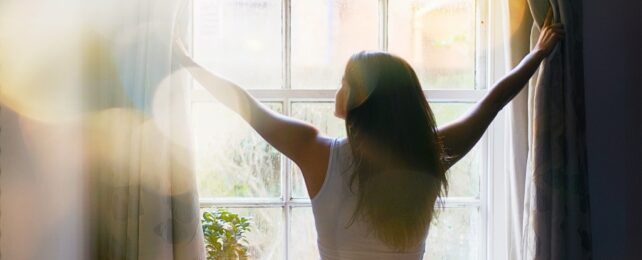 The back of a women with long brown hair opening curtains to reveal sun