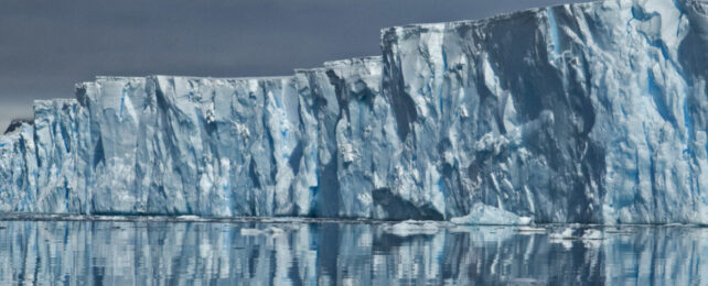 photo of an immense blue-white ice cliff towering over still ocean water.