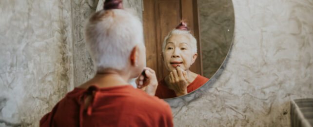 Asian woman with white hair wearing red sweater applies lipstick while looking in mirror.