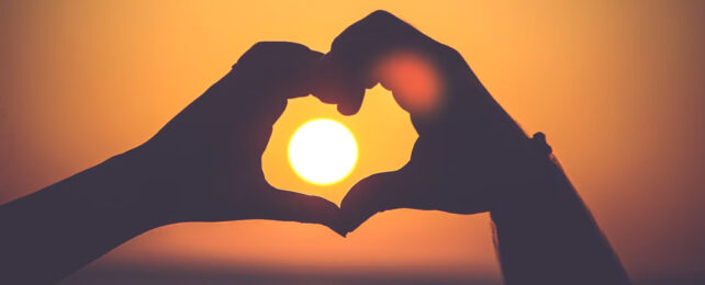 Silhouette of two people's hands forming a heart around the setting sun