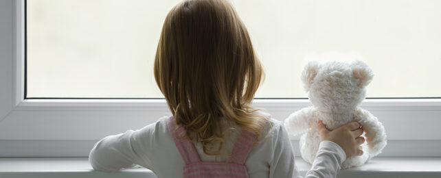 girl staring out a window while holding a teddy bear