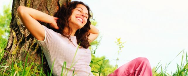 A smiling person with closed eyes sitting against a tree