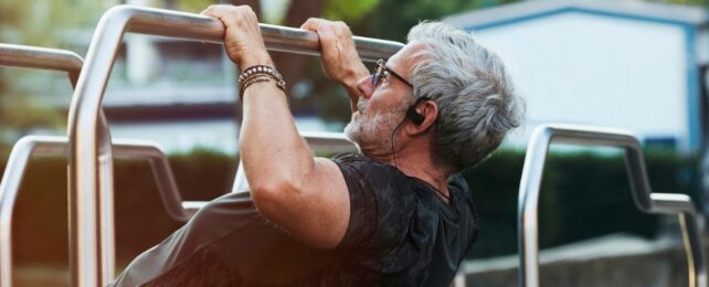 A man with grey hair wearing a black t-shirt doing a pullup on a bar outdoors