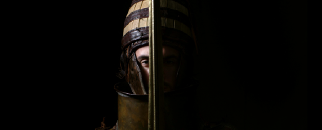 Close up of person wearing bronze armour holding sword in front of their face.