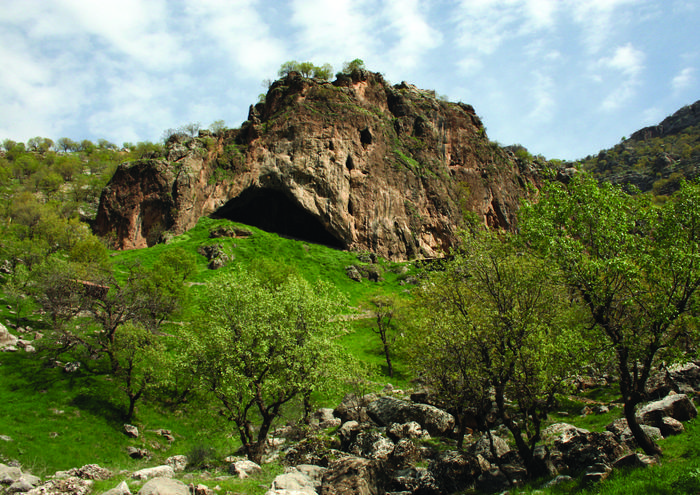 Entrance of a cave at the base of a rocky outcrop surrounded by shrubs and grass