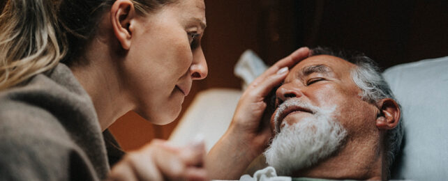 woman looking on with concern at an old man laying in bed with his eyes closed