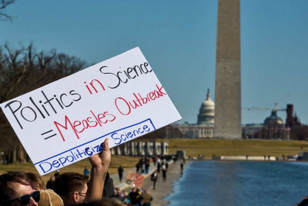a person's hand holds up a sign saying politics in science = measles outbreak, depoliticise science, in front of the US capitol building