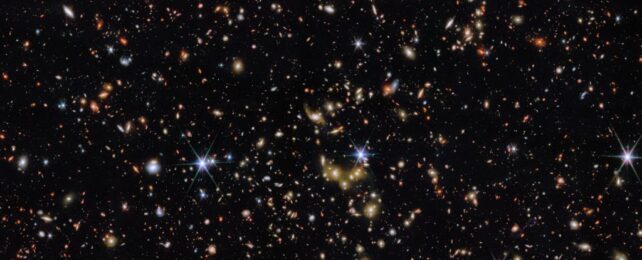 An area of deep space with thousands of galaxies in various shapes and sizes on a black background. Most are circles or ovals, with a few spirals. More distant galaxies are redder in colour and smaller, down to being mere dots, while closer galaxies are a bit larger and white or blueish. A few gold-coloured galaxies are bunched closely together in the centre. Bright stars surrounded by spikes lie in our galaxy.