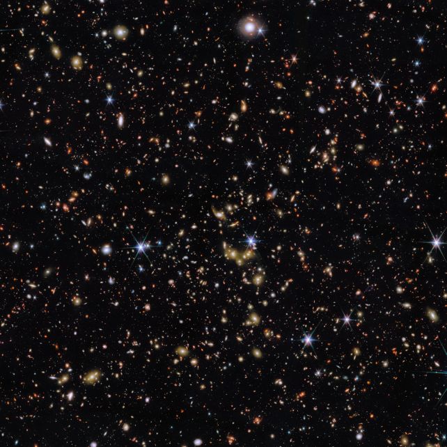 An area of deep space with thousands of galaxies in various shapes and sizes on a black background. Most are circles or ovals, with a few spirals. More distant galaxies are redder in colour and smaller, down to being mere dots, while closer galaxies are a bit larger and white or blueish. A few gold-coloured galaxies are bunched closely together in the centre. Bright stars surrounded by spikes lie in our galaxy.