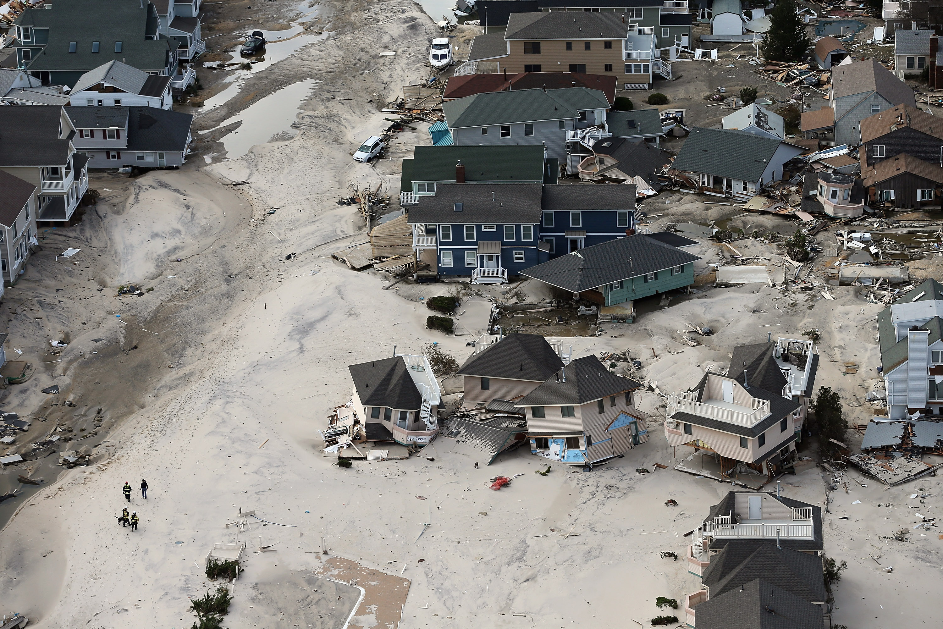 aerial photo of an american suburban neighbourhood destroyed by the storm. many grey-roofed houses are askew and destroyed, streets flooded with mud, a few people visible in the bottom left