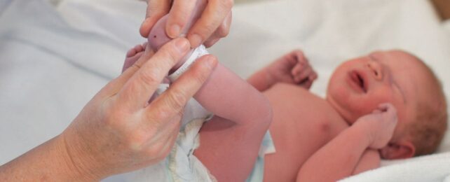 a newborn baby crying with foot in the air held by an adult hand, pinching the baby's heel which has a small drop of blood on it