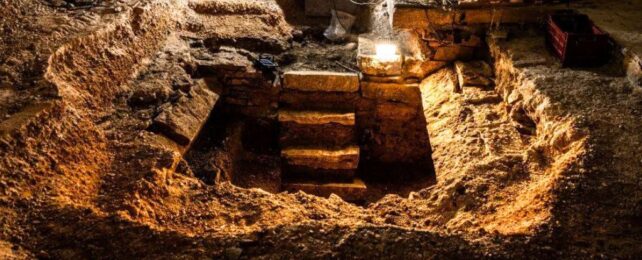 photo of an old dark dirt and stone floor, dug up to reveal a square hole with a stone staircase leading into it.