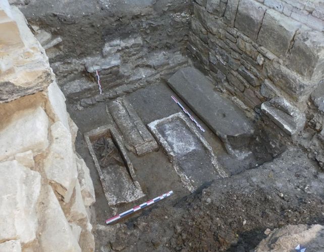 high angle shot of a rectangular depression in stone foundations revealing four sarcophagi, one containing visible human bones.