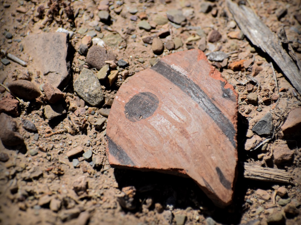 photo of a piece of terra cotta ceramic, with some dark lines and a dot painted on, on a gravel ground