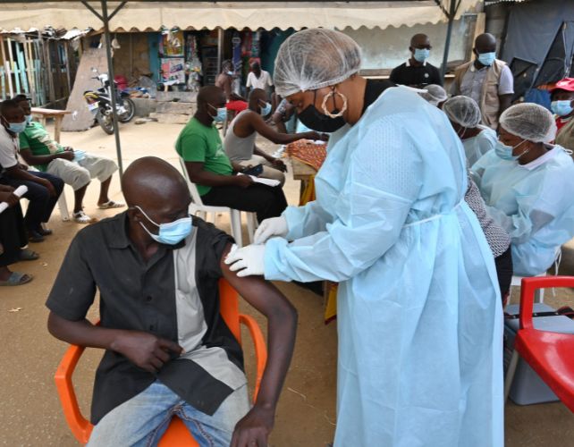 A health worker vaccinates a man who is sitting in a plastic chair. they are under an outdoor marquee, with many other peoples sitting around behind them. everyone is wearing face masks.