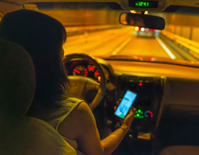 a photo taken over the shoulder of a young woman driver who is holding her phone while driving at night.