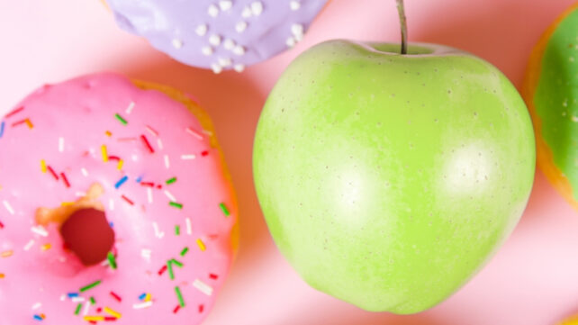 Close up of green apple next to pink donut on a pink background.