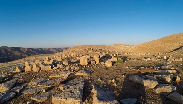 A collection of oblong shaped stones arranged loosely in lines, on top of a knoll with no vegetation. Nearby hills are in the background and the sky is blue.