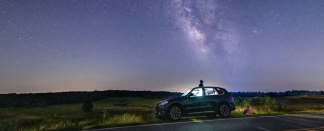 a car parked on the side of the road in a remote field. the milky way and a meteor are visible in the night sky. a person sits on top of the car, another person sits beside it, both looking at the sky.