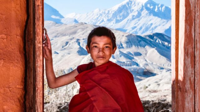A young monk stands in an open doorway, with snowy mountains in the background