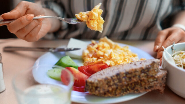 Person in a striped shirt is sitting at a table eating breakfast, holding up scrambled eggs on their fork