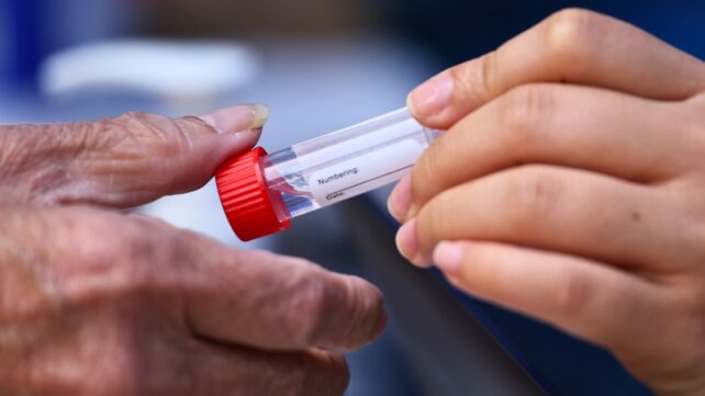 a young adult's hand passing over a saliva sample tube to an older person's hand